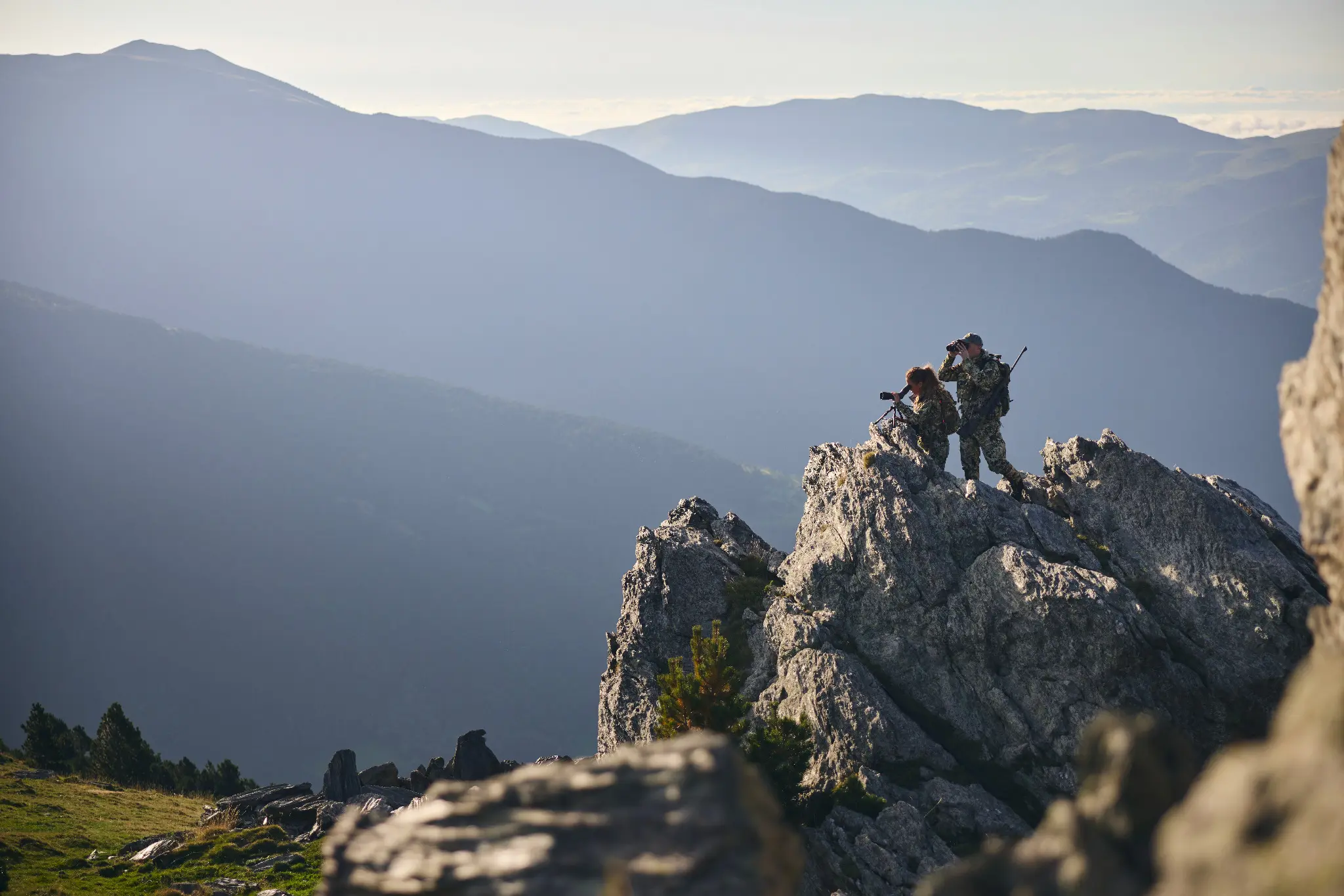 Two people using optics to observe a mountain landscape from a rocky ridge